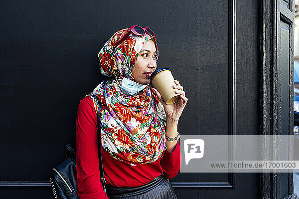 Muslim woman while drinking coffee against black wall during COVID-19