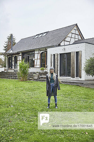 Ältere Frau in Regenmantel steht im Hinterhof vor einem Haus
