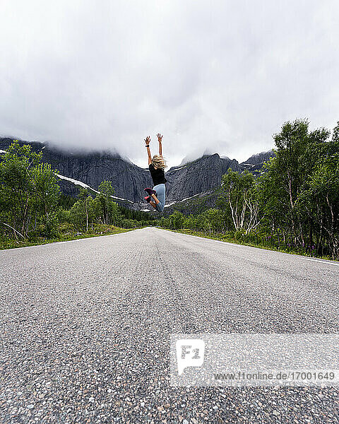 Frau mit erhobener Hand springt auf der Straße gegen einen Berg auf den Lofoten  Norwegen