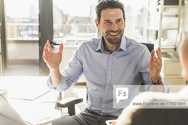 Lächelnder Geschäftsmann im Gespräch mit einem Kollegen  während er im Büro sitzt