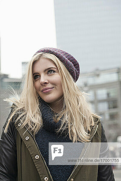 Lächelnde blonde Frau mit Strickmütze schaut weg  während sie in der Stadt steht