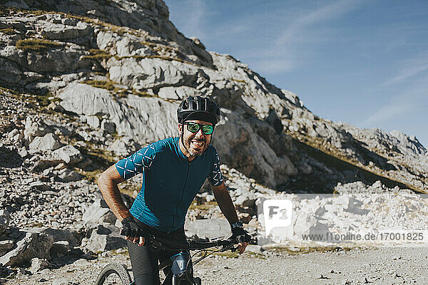 Fröhlicher männlicher Radfahrer auf dem Fahrrad gegen einen Berg an einem sonnigen Tag  Nationalpark Picos de Europa  Kantabrien  Spanien