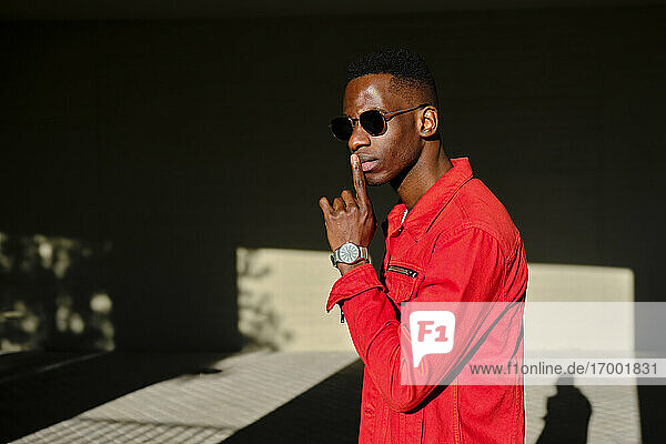 Young man in red jacket and sunglasses with finger on lips outdoors