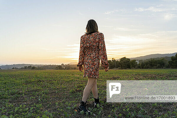 Junge Frau in geblümtem Sommerkleid geht bei Sonnenuntergang auf einer Wiese spazieren