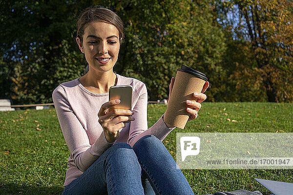 Lächelnde Unternehmerin  die ein Smartphone benutzt  während sie mit einer Kaffeetasse im Herbst im Park sitzt