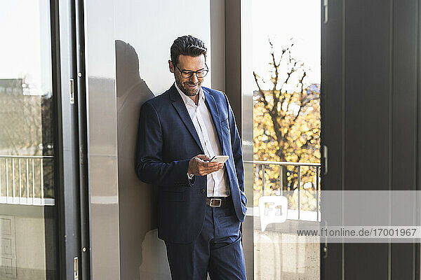 Männlicher Unternehmer  der ein Mobiltelefon benutzt  während er mit der Hand in der Tasche auf dem Balkon eines Büros steht