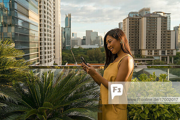 Lächelnde Frau steht auf einer Dachterrasse und schaut auf ihr Smartphone  Bangkok  Thailand