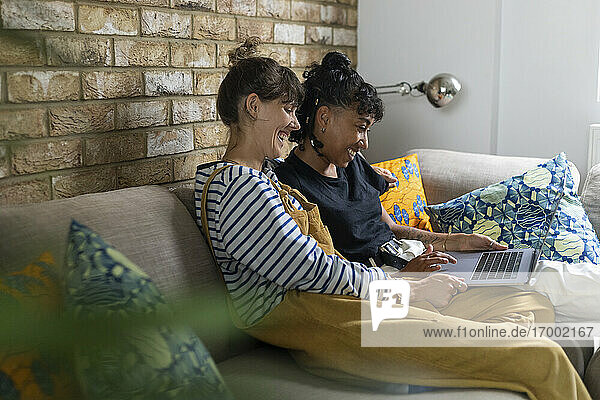 Lesbisches Paar  das lächelnd einen Laptop benutzt und zu Hause auf dem Sofa sitzt