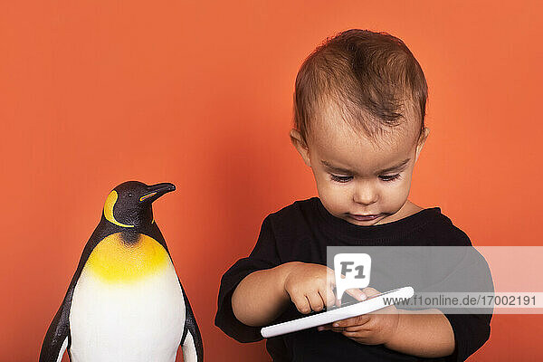 Kleinkind ignoriert Pinguin-Spielzeug  während es ein Mobiltelefon vor orangefarbenem Hintergrund benutzt