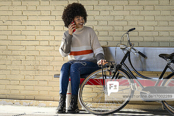 Happy female passenger talking through mobile phone while sitting on bench by bicycle against brick wall