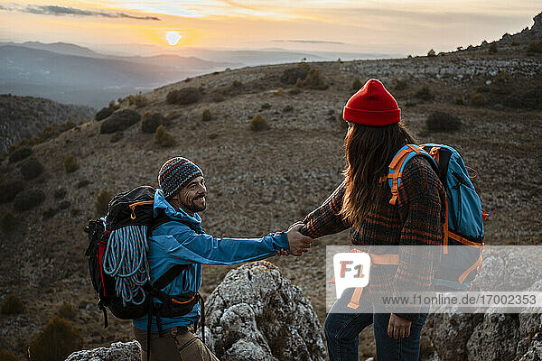 Freundin hilft ihrem Freund beim Klettern auf einen felsigen Berg bei Sonnenuntergang
