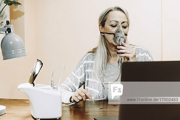 Ältere Frau mit geschlossenen Augen bei der Verwendung eines Verneblers während einer Online-Beratung zu Hause