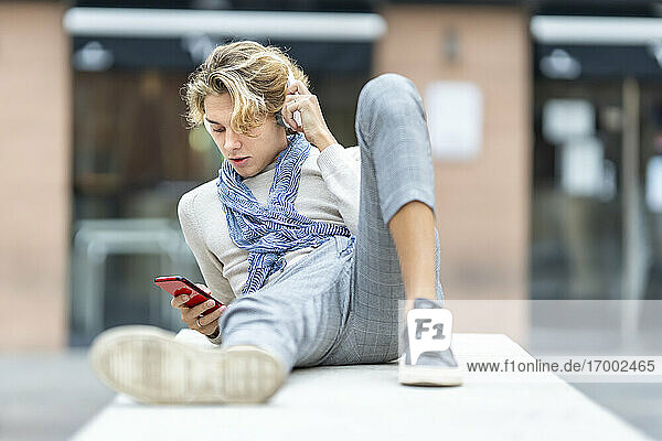 Junger Mann mit Kopfhörern  der ein Mobiltelefon benutzt  während er sich auf einer Bank in der Stadt ausruht