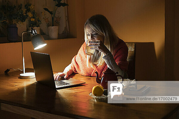 Frau trinkt Tee  während sie einem Videoanruf zuhört  während sie zu Hause sitzt