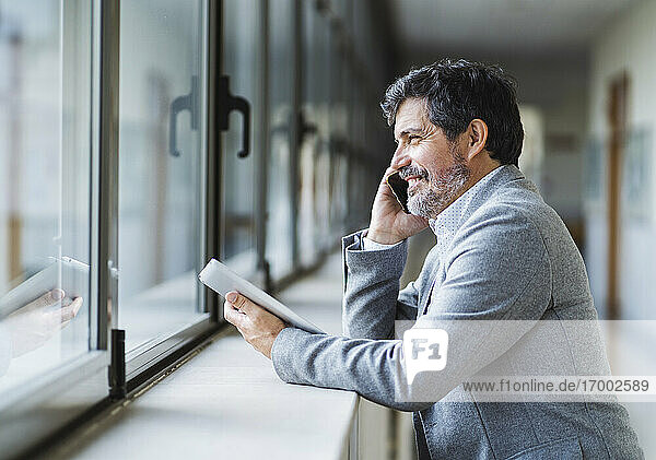 Lächelnder männlicher Professor  der am Telefon spricht und durch ein Fenster im Korridor einer Universität schaut