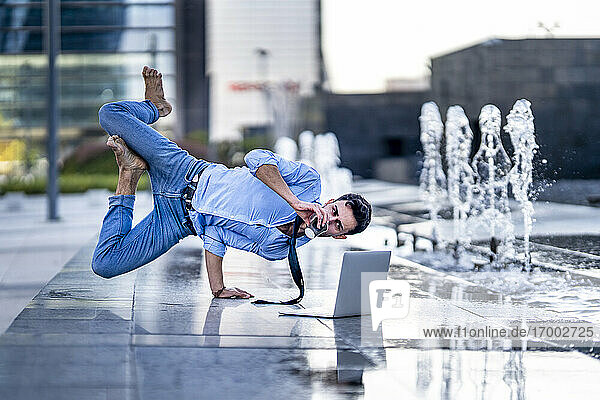 Businessman with laptop drinking coffee while doing handstand by fountain on footpath