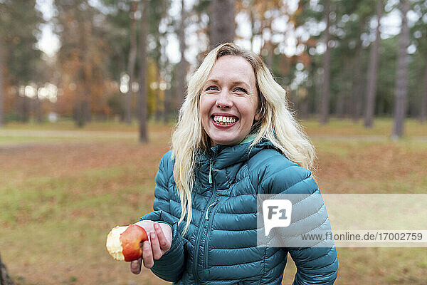 Glückliche blondhaarige Frau hält einen Apfel in der Hand  während sie in Cannock Chase steht