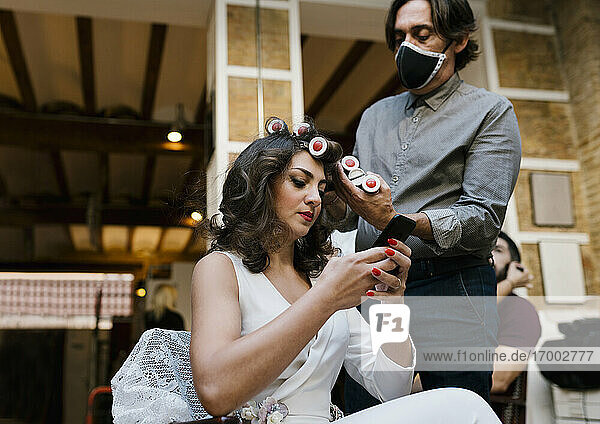 Männlicher Friseur  der eine Frisur macht  während die Braut ihr Smartphone in einem Salon während einer Pandemie benutzt