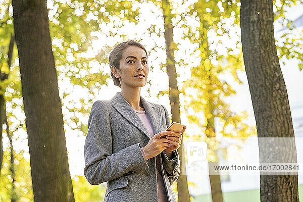 Weibliche Unternehmerin  die wegschaut  während sie ihr Smartphone in einem Park im Herbst hält
