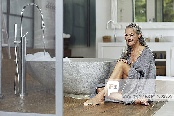 Lächelnde reife Frau sitzt bei der Badewanne auf dem Boden im Badezimmer