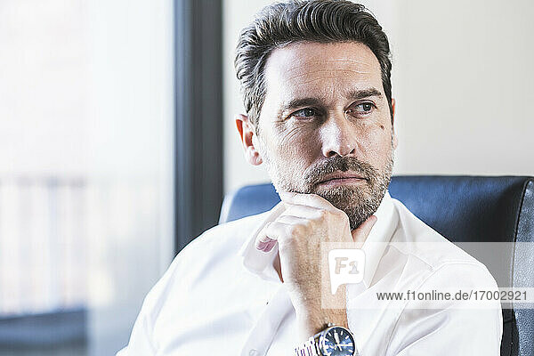Businessman with hand on chin looking away while sitting at office