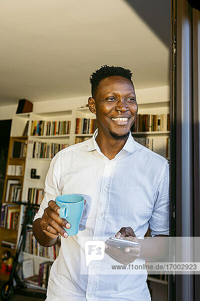 Glücklicher Geschäftsmann mit Kaffeetasse und Smartphone in der Hand  während er sich zu Hause an die Tür lehnt