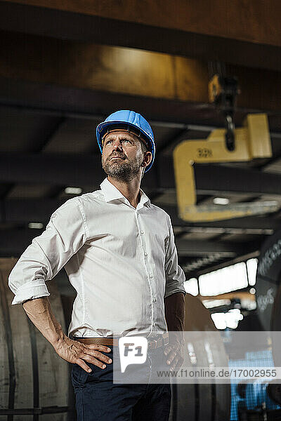 Businessman with hands on hip looking up while standing in factory