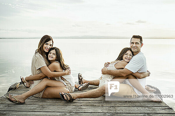 Daughters embracing parents while sitting on jetty by lake against sky