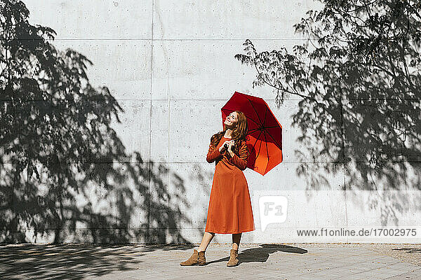 Lächelnde Frau  die einen Regenschirm hält  während sie vor einer Baumschattenwand steht