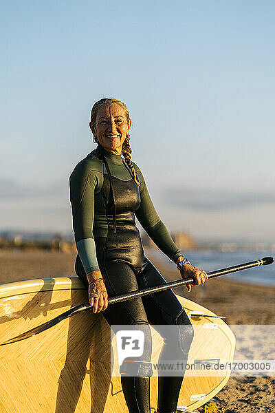 Lächelnde Frau  die sich in der Morgendämmerung am Strand auf ein Paddleboard stützt