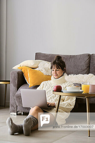 Frau im Pullover  die einen Laptop benutzt  während sie im Wohnzimmer zu Hause auf dem Boden sitzt