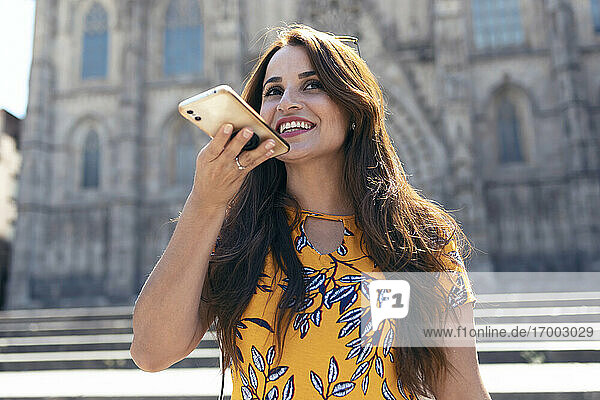 Lächelnde erwachsene Frau  die mit einem Mobiltelefon spricht  während sie am Platz der Kathedrale von Barcelona in Barcelona  Katalonien  Spanien  steht