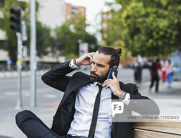 Portrait of bearded businessman sitting on bench and talking on smart phone