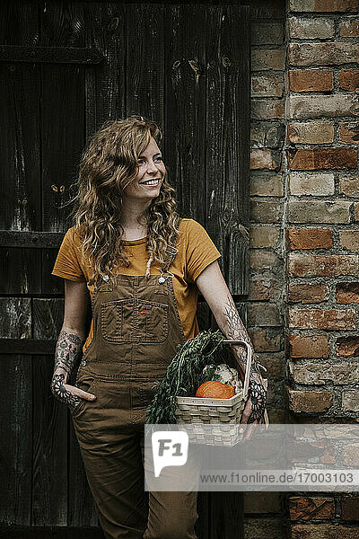 Lächelnde Frau hält Gemüsekorb gegen Wand