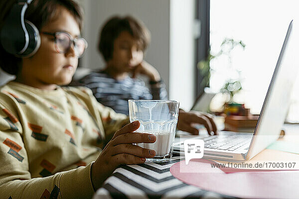 Junge hält ein Glas Milch in der Hand  während er zu Hause einen Laptop benutzt