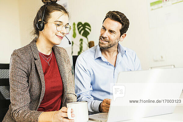 Lächelnde Geschäftsfrau mit Kopfhörern  die Kaffee trinkt  während sie mit einem Kollegen am Laptop im Büro arbeitet