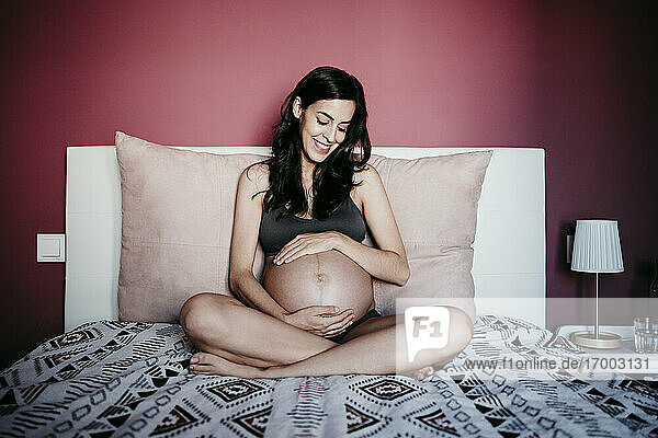 Glückliche schwangere Frau  die ihren Bauch betrachtet  während sie im Schlafzimmer zu Hause sitzt