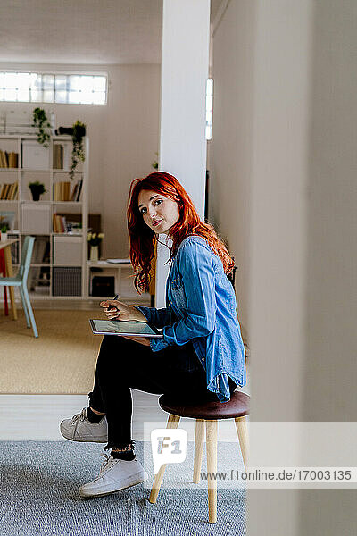 Rothaarige Geschäftsfrau  die ein digitales Tablet benutzt  während sie auf einem Hocker im Büro sitzt