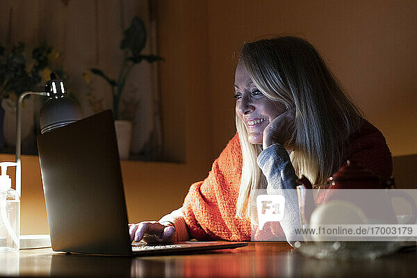 Lächelnde Frau  die einen Videoanruf auf dem Laptop hört  während sie zu Hause am Tisch sitzt