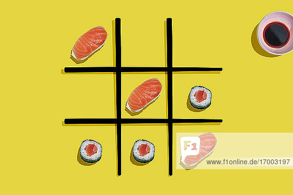 Tic-tac-toe game with salmon sushi maki and nigiri with black chopsticks on yellow background