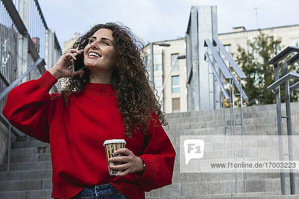 Lächelnde Frau  die eine Kaffeetasse hält  während sie auf einer Treppe mit ihrem Handy spricht