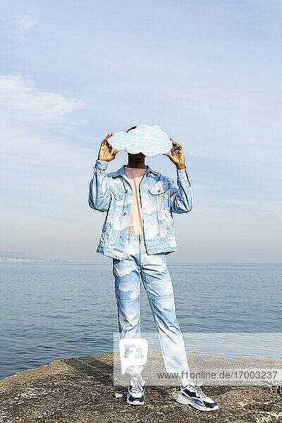 Junger Mann versteckt Gesicht mit ausgeschnittener Wolke  während er am Meer steht