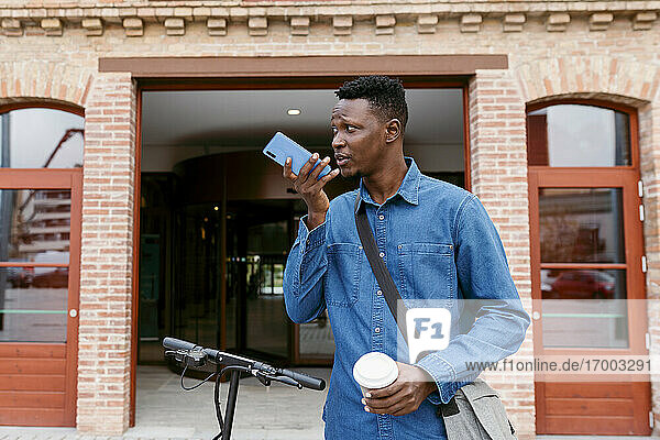 Männlicher Unternehmer  der eine Voicemail über sein Smartphone sendet  während er vor einem Gebäudeeingang steht