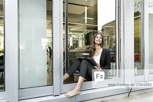 Lächelnde Geschäftsfrau mit digitalem Tablet  die wegschaut  während sie sich an die Bürotür lehnt