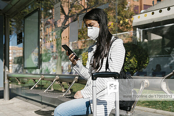 Frau wartet auf den Bus und benutzt ihr Smartphone an einem sonnigen Tag während der COVID-19-Pandemie