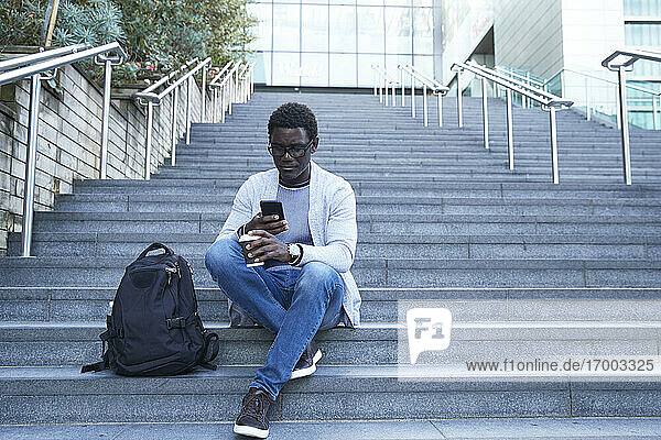 Unternehmer  der ein Mobiltelefon benutzt  während er einen Einwegbecher auf einer Treppe im Finanzviertel hält