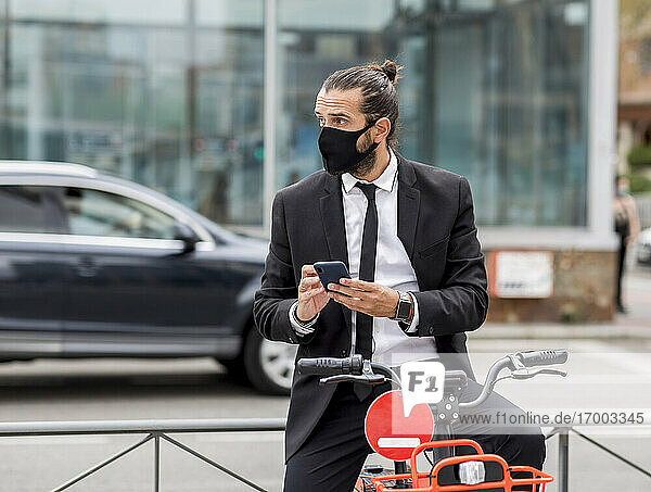 Portrait of businessman wearing protective face mask sitting on bicycle with smart phone in hands