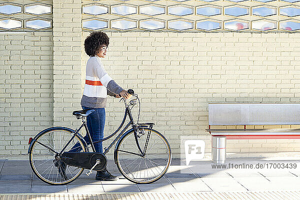 Smiling young woman wheeling bicycle by brick wall at station