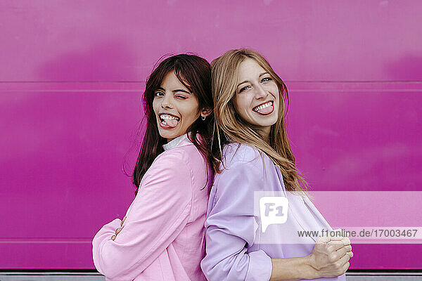 Schwestern strecken die Zunge heraus  während sie Rücken an Rücken an einer rosa Wand stehen