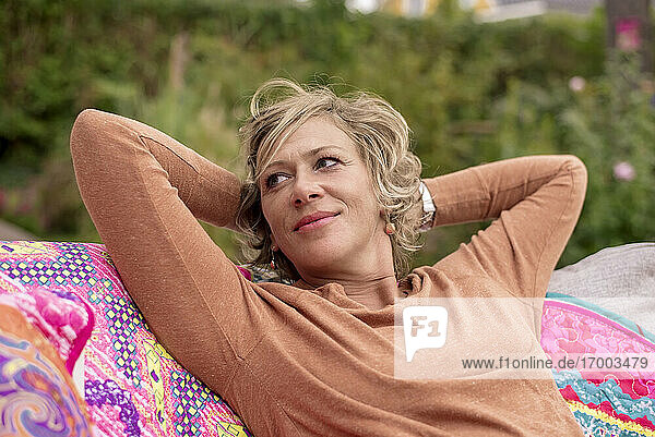 Lächelnde Frau sitzt mit den Händen hinter dem Kopf auf einem Sofa im Hinterhof